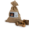 Ein Jutesack mit Olivenholz-BBQ-Chunks, der mit einer Schnur oben verschlossen ist. Auf dem Sack befindet sich ein Etikett mit Produktinformationen und einem Bild von brennenden Holzstücken. Vor dem Sack liegen mehrere Stücke Olivenholz in unterschiedlichen Formen und Größen, die eine natürliche, rustikale Optik haben.